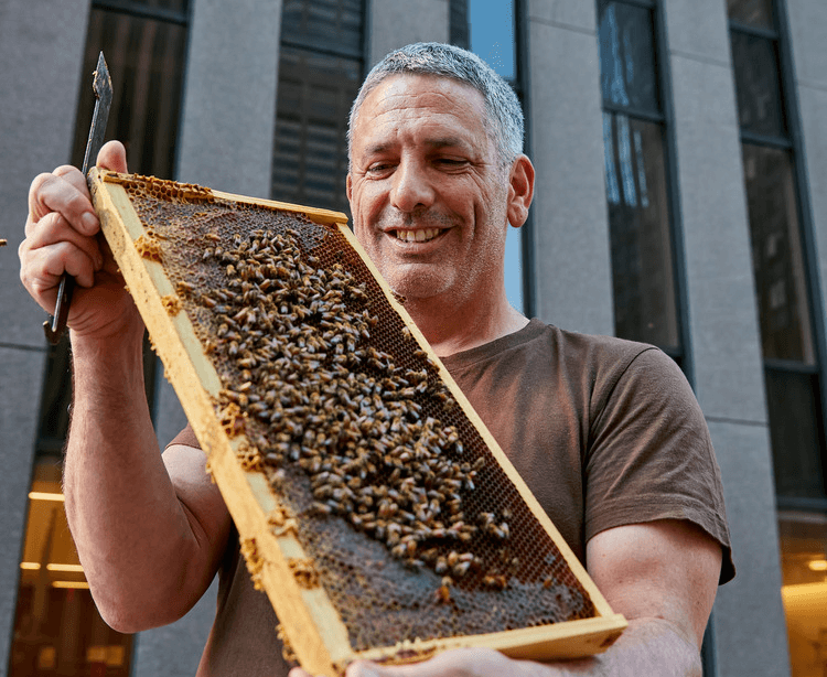 Man in brown shirt smiling while holding bee frame with honeycomb full of bees outside building.