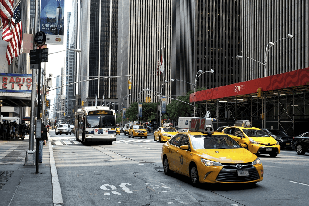 Yellow taxis and a city bus drive down a busy street in Manhattan, New York City, with tall buildings and Radio City Music Hall visible.