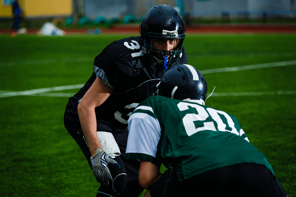 Two American football players face each other on a field; one in a black uniform with number 31, the other in green with number 20, preparing for a play.